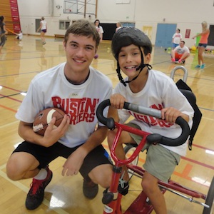 An older boy holding a ball and smiling next to younger boy on a tricycle